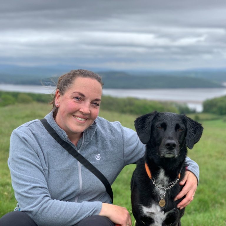 Woman smiling beside a black dog Woman smiling beside a black dog, with a scenic landscape in the background.